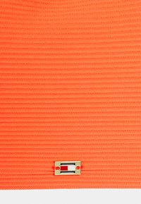 Tecido texturizado laranja brilhante com riscas horizontais, apresentando um logótipo quadrado metálico na parte inferior.