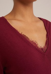 Maglione bordeaux a V con delicata rifinitura in pizzo sul collo; texture morbida e dettagli frangiati sui bordi. Vista ravvicinata.