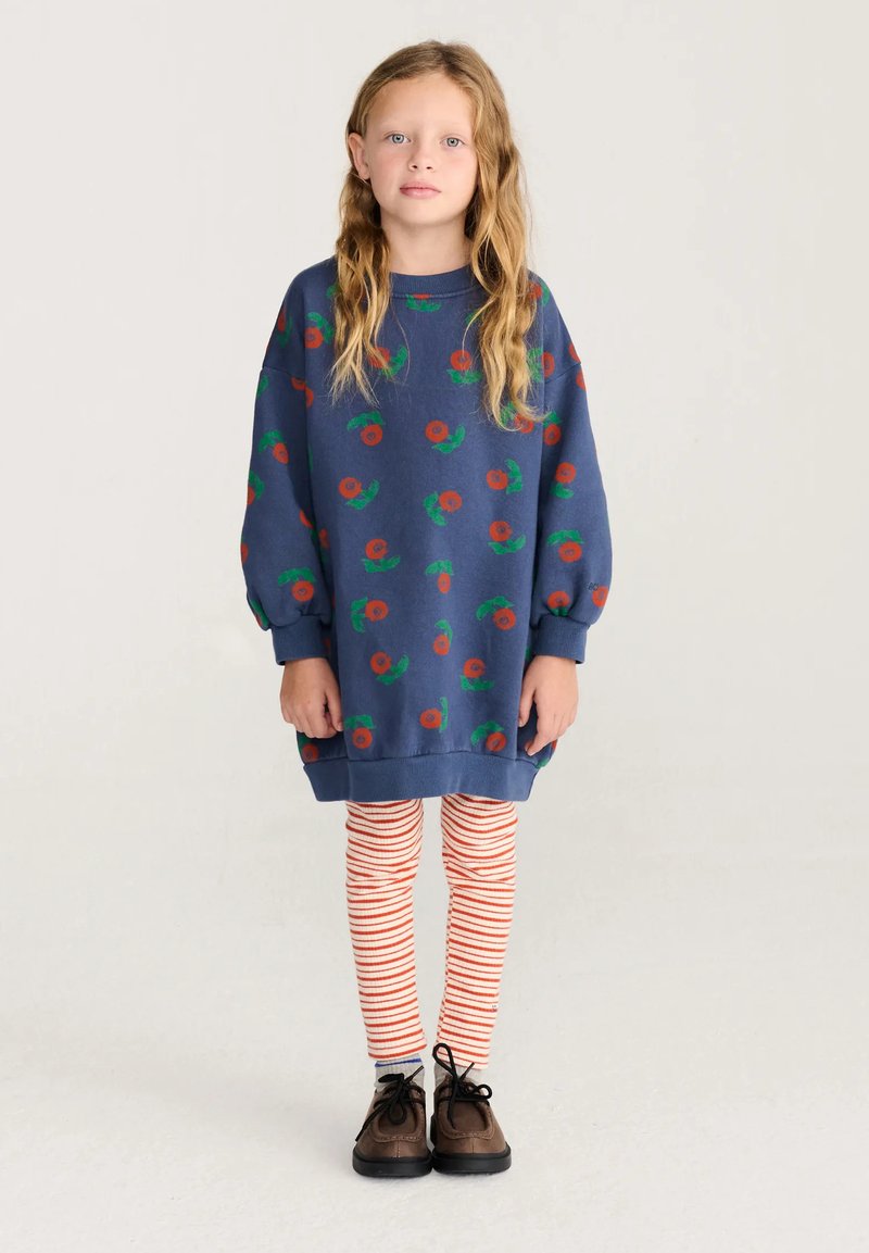 Jeune fille aux longs cheveux blonds portant un pull bleu oversize avec des fleurs rouges, des leggings rayés et des chaussures marron, debout devant un fond uni.