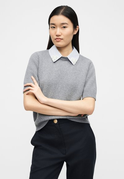 Femme aux cheveux noirs raides portant un pull gris à manches courtes par-dessus une chemise rayée à col avec broderie florale, assorti à un pantalon noir.