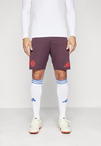 Pantalones cortos atléticos burdeos con el logo rojo del Bayern de Múnich, combinados con calcetines blancos con rayas azules y zapatillas blancas con acentos naranjas.