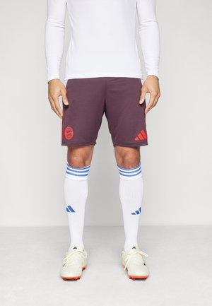 Pantalones cortos atléticos burdeos con el logo rojo del Bayern de Múnich, combinados con calcetines blancos con rayas azules y zapatillas blancas con acentos naranjas.