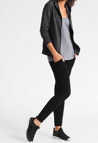 Sudadera con capucha negra con cremallera, camiseta de tirantes gris, pantalones jogger ajustados negros y zapatillas deportivas negras. Tejido suave, ajuste relajado y diseño minimalista.