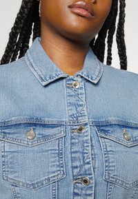Světlomodrá džínová bunda s klasickým límcem, stříbrnými cvoky a dvěma náprsními kapsami s klopami. Texturovaná tkanina a detaily šití.