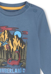 Camicia a maniche lunghe blu con una grafica colorata di un paesaggio all'aperto che presenta montagne, alberi e una creatura rossa, impreziosita da dettagli gialli nel collo.