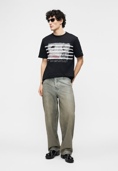 Jeune homme portant un t-shirt noir à motifs, un jean gris oversize, des chaussures noires et des lunettes de soleil foncées, debout les mains dans les poches.