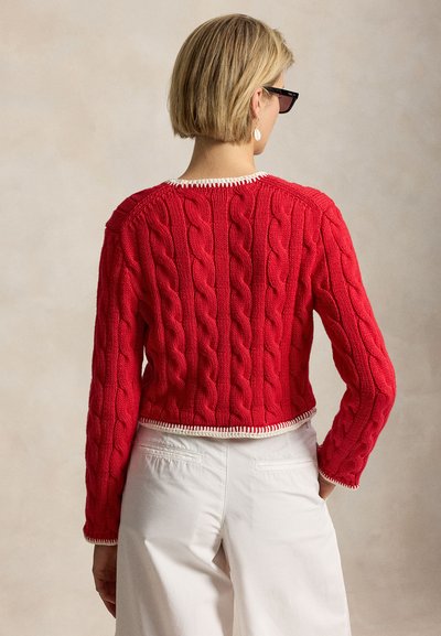 Kobieta o krótkich blond włosach, w czarnych okularach przeciwsłonecznych, czerwonym swetrze w warkocz z białym wykończeniem i białych spodniach, zwrócona tyłem do kamery.