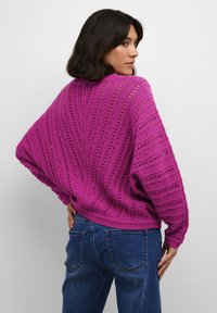 Maglione over-size fucsia con motivo di tricot aperto a zigzag, polsini e orlo a coste, abbinato a jeans blu a vita alta.