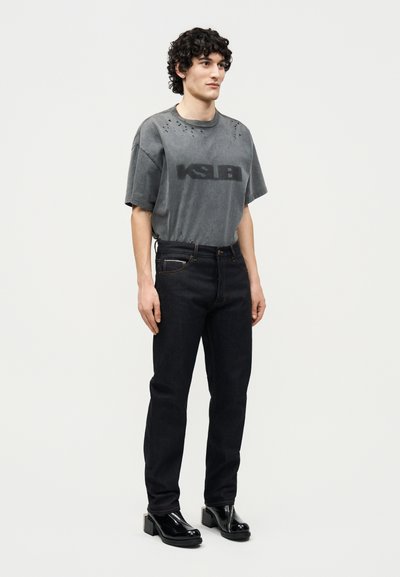 Νέος άνδρας με σγουρά μαλλιά φορώντας μία ξεθωριασμένη μαύρη φαρδιά μπλούζα KSUBI, μαύρα ίσια τζιν και γυαλιστερά μαύρα παπούτσια με πλατφόρμα.