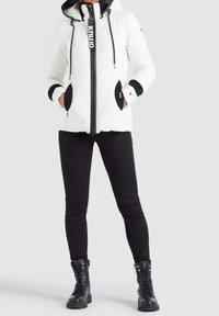 Veste matelassée blanche avec des accents noirs, capuche, cordons de serrage et fermeture éclair. Poches noires et poignets côtelés. Associée avec un pantalon noir et des bottes.