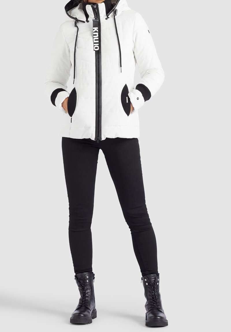 Veste matelassée blanche avec des accents noirs, capuche, cordons de serrage et fermeture éclair. Poches noires et poignets côtelés. Associée avec un pantalon noir et des bottes.