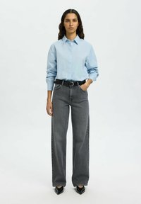 Camicetta azzurro chiaro con maniche lunghe, abbinata a jeans grigi scuri a vita alta e a gamba larga, e scarpe col tacco nere a punta.