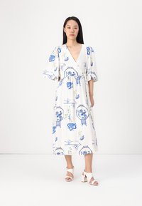 Weißes Wickelkleid mit blauem Blumenmuster, kurzen Puffärmeln und einem Bindeband in der Taille. Modell trägt es mit weißen Blockabsatzsandalen.