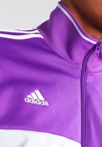Lila zip-jacka med hög krage, vita kontrastlinjer och broderad Adidas-logotyp i vitt på vänster bröst. Slät tygtextur.