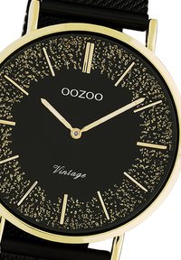 Montre noire avec des accents dorés, cadran rond, cadran texturé avec paillettes dorées, marqueurs d'heure avec des lignes fines et un bracelet noir.