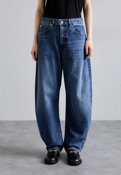 Weite blaue Denim-Jeans mit hoher Taille, klassischem Fünf-Taschen-Design und verblasstem Textur, kombiniert mit schwarzem Schuhwerk und goldenen Akzenten.