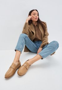 Mocassini in pelle tan con punta arrotondata e un'applicazione in metallo dorato. Texture liscia con cuciture minime, abbinati a jeans azzurri.
