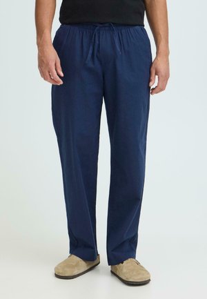 Mand iført marineblå bukser med snøre og beige slip-on sko, stående mod en ensartet lys baggrund.