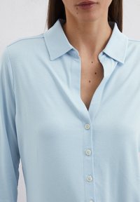 Light blue button-up shirt with a soft texture, featuring a collared neckline and seven front buttons.