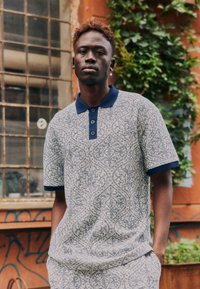 Jeune homme aux cheveux courts et bouclés portant un polo à manches courtes à motifs et un pantalon assorti, debout à l'extérieur près de graffitis et de verdure.
