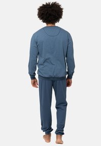 Henkilö, jolla on kiharat hiukset, pukeutuneena siniseen kuvioituun pitkähiharaiseen puseroon ja yksivärisiin sinisiin housuihin, seisoo paljain jaloin selkä kameralle päin yksinkertaisella taustalla.