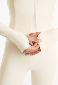 Body aderente a maniche lunghe in morbido tessuto beige chiaro a coste, con zip frontale e aperture per il pollice sui polsini, che mette in risalto la texture.