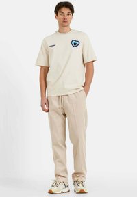Beige katoenen t-shirt met blauwe hartborduursels, gecombineerd met beige drawstring broeken en lichte sneakers met zwarte accenten en rubberen zolen.