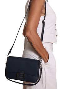 Vrouw die een witte mouwloze top en broek draagt, met een zwarte leren schoudertas en een tattoo op haar onderarm.