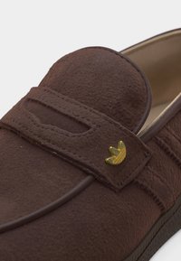 Κοντινή λήψη ενός καφέ σουέτ μοκασίνι με ιμάντα που έχει ένα μικρό χρυσό έμβλημα στο πλάι.