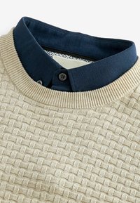Pull en maille beige texturée superposé sur un col de chemise bleu foncé boutonné.