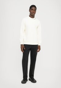 Young man standing, wearing a white sweatshirt with logo on the collar, black pants, and black sneakers against a plain light background.