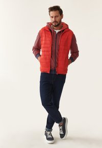 Rød puffervest over en marineblå og rød stribet hættetrøje, parret med mørke denimjeans og grå sneakers med hvide såler og snørebånd.