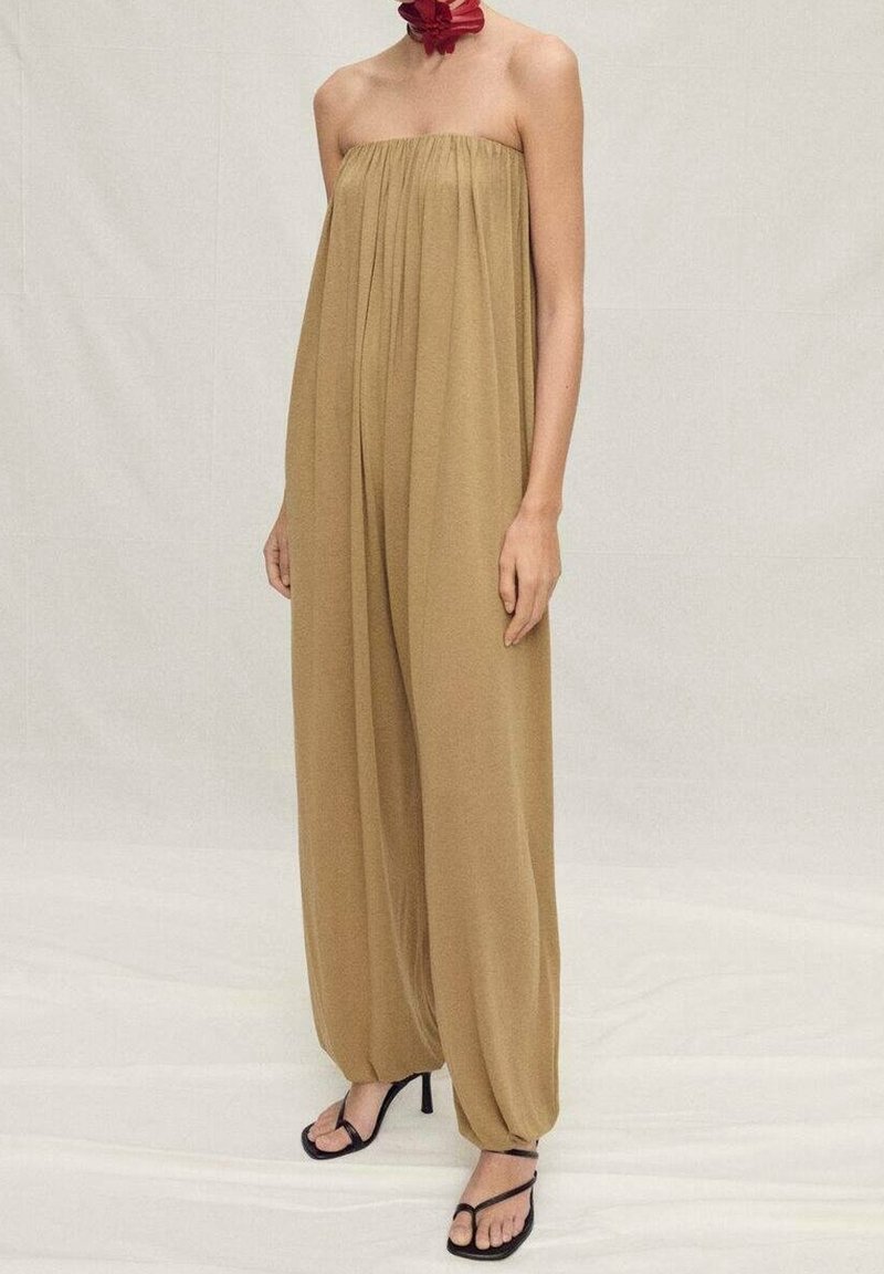 Femme portant une combinaison beige bustier à jambes larges et des sandales à talons noires, debout sur un fond clair uni.