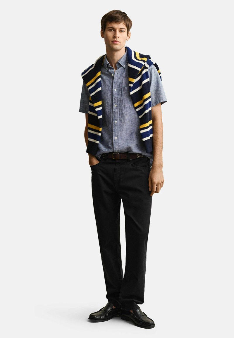 Giovane uomo in piedi che indossa una camicia blu a maniche corte con bottoni, pantaloni neri, scarpe nere e un maglione a righe drappeggiato sulle spalle.