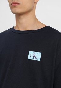 Marinblå bomulls-t-shirt med rund halsringning, som har en ljusblå rektangulär logotyp-etikett med texten "Calvin Klein Jeans" i lila.