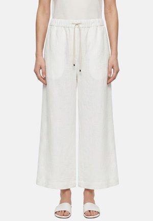 Donna che indossa pantaloni larghi in lino bianco con coulisse in vita e sandali bianchi scivolati, in piedi davanti a uno sfondo uniforme.