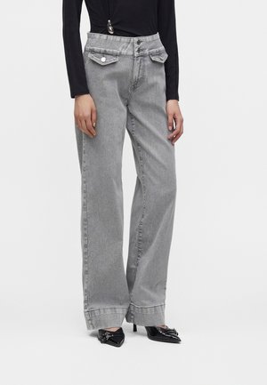 Personne portant un jean gris taille haute à jambes larges avec fermeture à double bouton et poches à rabat, associé à des talons noirs pointus.