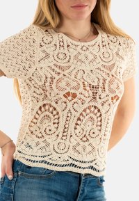 Top de ganchillo en beige con un diseño de calado detallado y patrones intrincados, mangas cortas y un dobladillo con volantes.