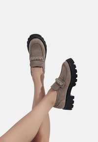 Mocassini in suede taupe con una suola in gomma nera chunky a contrasto. Presentano un dettaglio a tracolla intrecciata sulla tomaia.