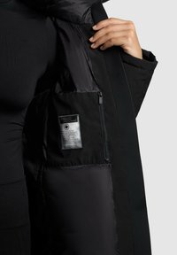 Eine Person in schwarzer Kleidung öffnet ihren schwarzen Mantel, um eine innere Reißverschlusstasche und ein rechteckiges Markenlabel im Innenfutter der Jacke zu zeigen.