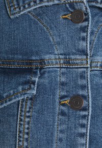 Giacca di jeans di un blu medio con cuciture gialle, dotata di bottoni metallici, tasche a toppa e design classico del colletto. Dettagli del tessuto testurizzato visibili.
