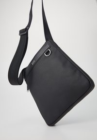 Black triangular shoulder bag made of textured fabric, featuring a zippered closure, adjustable strap, and a metal D-ring accent.
