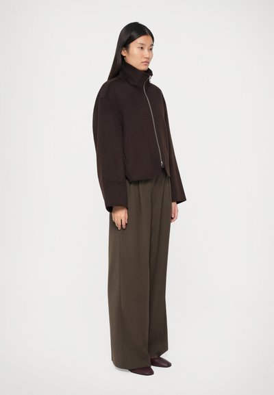 Jachetă scurtă maro cu guler înalt și fermoar frontal, purtată cu pantaloni largi de culoare verde măsliniu și pantofi negri. Textură netedă.