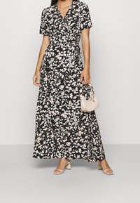 Czarna sukienka maxi w kwiaty z krótkimi rękawami i designem typu wrap, z delikatnymi akcentami w kolorze brzoskwiniowym i białym. Stylizowana z małą beżową torebką.
