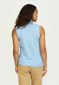 Top brez rokavov iz modrega pletenega materiala z visokim ovratnikom, gladko teksturo in rebrastim robom. Uporabljen s svetlim bež hlačami za sproščen videz.