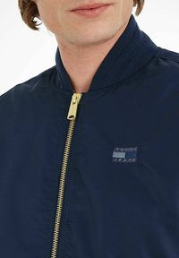 Giubbotto bomber blu navy con zip dorata, polsini a coste e un piccolo logo ricamato "TOMMY JEANS" in blu e bianco sul petto.