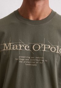 Olijfgroene katoenen t-shirt met geborduurde tekst van de merknaam "Marc O'Polo" en een slogan over natuurlijke erfgoed.