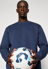 Sudadera azul marino con "CFC" y el logo de Nike en azul claro, sostenida sobre un balón de fútbol blanco con patrones geométricos azules y negros.