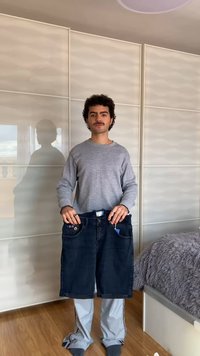 Jeune homme en pull gris et pantalon clair tenant un short en jean noir surdimensionné dans une chambre avec un lit et une armoire derrière lui.
