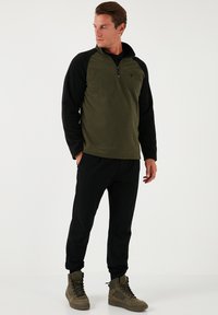 Olíva zöld testtel és fekete ujjal rendelkező pulóver, texturált fleece anyagból, cipzáras nyakkal, fekete joggingnadrággal és világosbarna magas szárú csizmával párosítva.